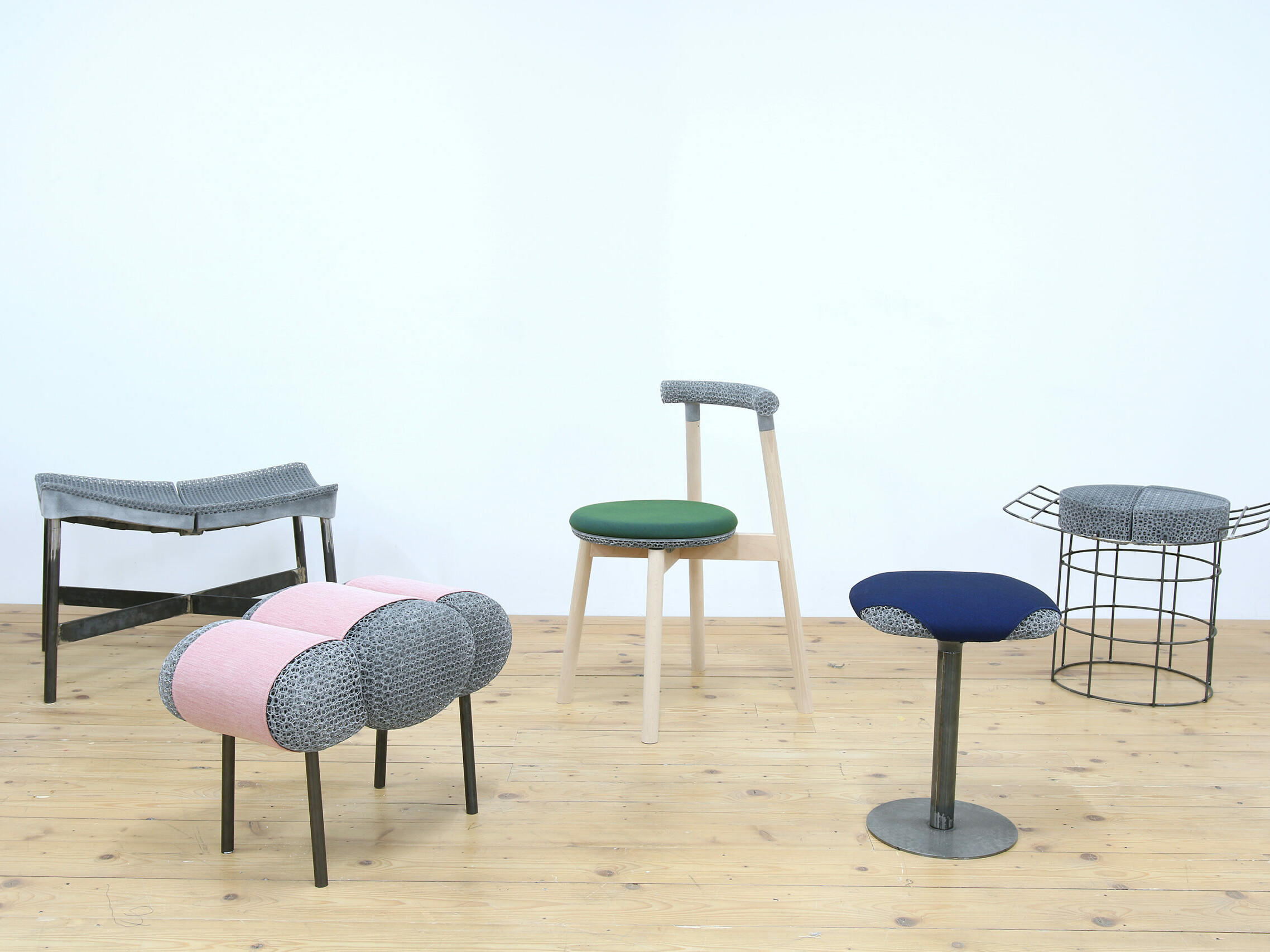 New possibilities of furniture production: Various chairs and stools in front of a white wall on a wooden floor with various substructures and 3D-printed seats and textile covers in blue, pink and green.