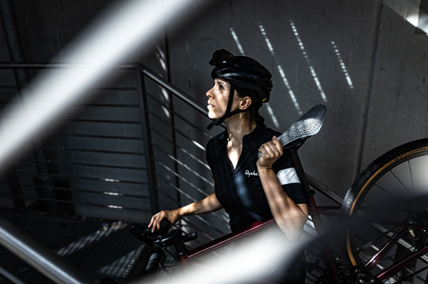 A white woman wearing a black shirt and helmet carries a bicycle with a 3d printed saddle as she climbs stairs in a stairwell with some sunlight streaming through.