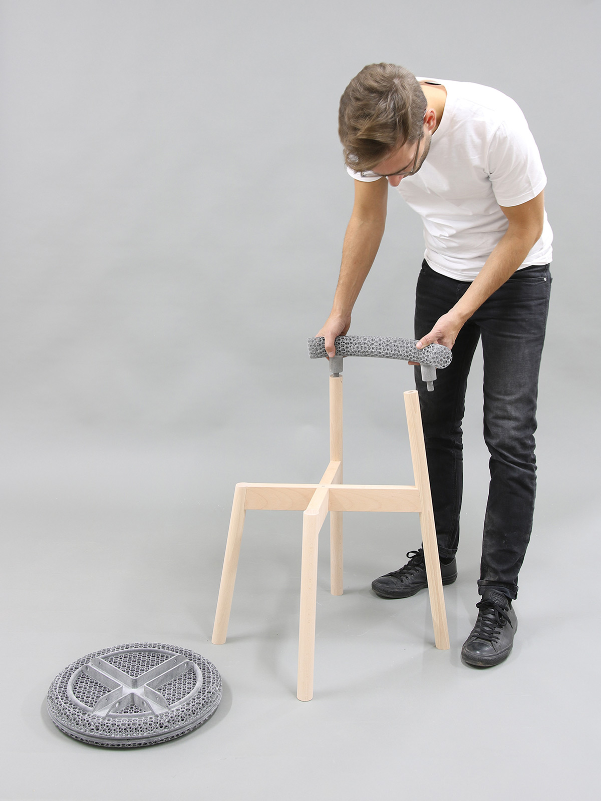 A white man with short hair and glasses shows how a chair with a wooden substructure and mesh seat can be disassembled separately.
