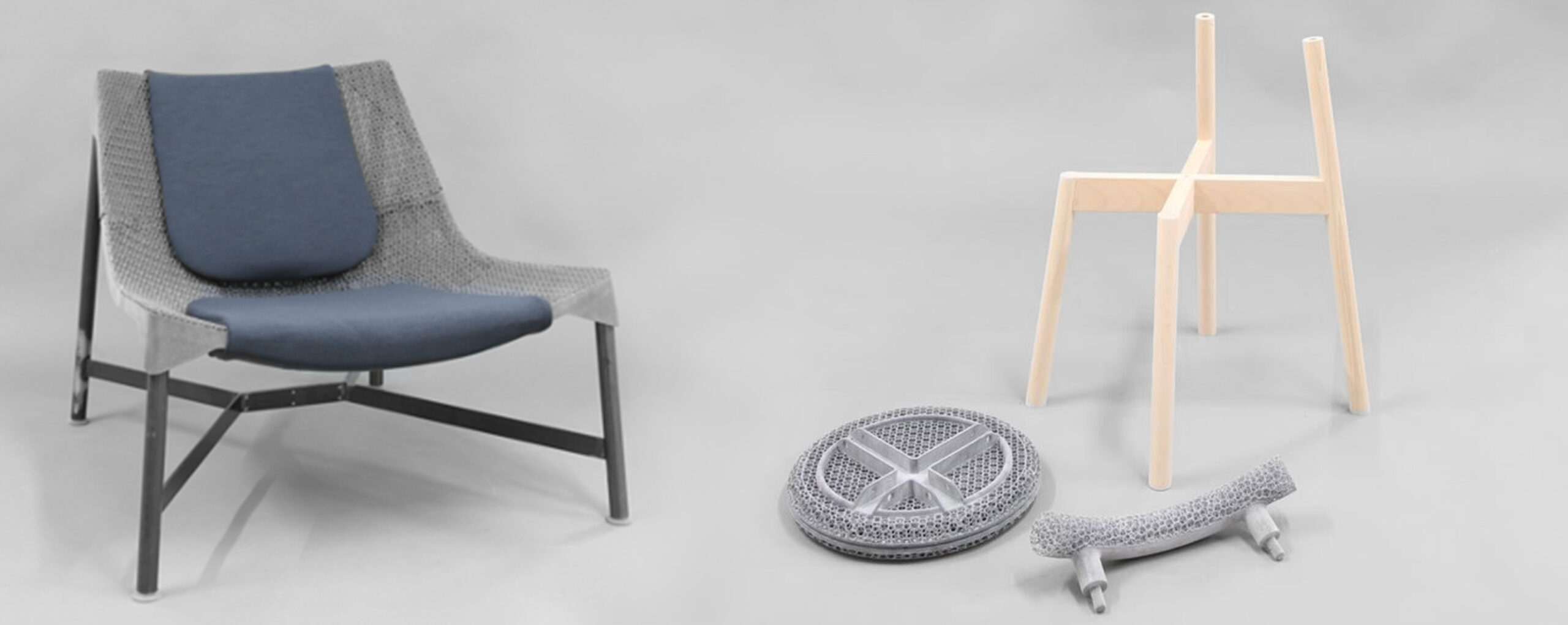 A chair with a metal substructure and a wide seat made of a gray lattice structure with dark gray seat cushions in the middle, next to it a wooden chair and a 3D-printed seat and backrest in individual parts lying on the floor.