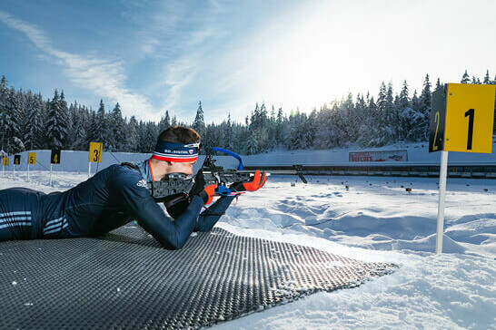 A young white biathlete surrounded by snow lying on the ground aiming his rifle at a target. Snow-covered fir trees in the background.