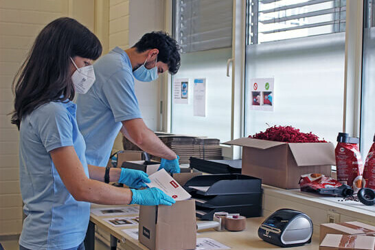 A young woman and a young man wearing protective masks and light blue polo shirts are packing small boxes while standing at a desk.