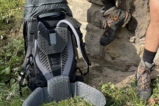 Feet of a white man in hiking boots on a rock, next to him the rear view of a backpack with grey lattice structure padding.