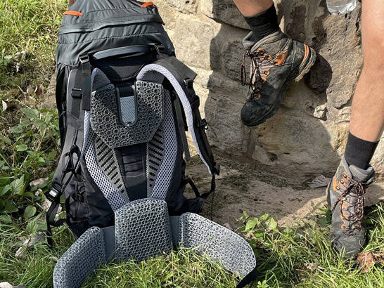 Feet of a white man in hiking boots on a rock, next to him the rear view of a backpack with grey lattice structure padding.