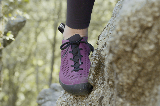 Close-up of a leg of a female rock climber standing with her foot on a rock wearing a purple black-soled shoe.