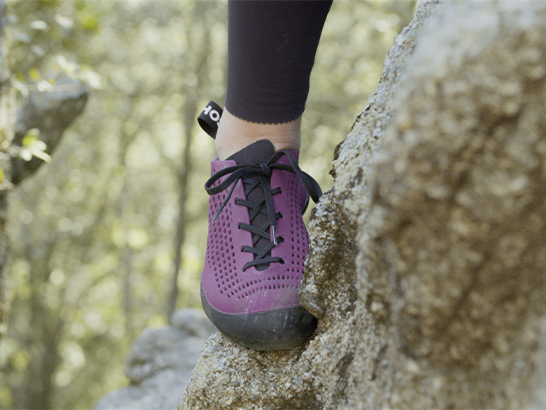 Close-up of a leg of a female rock climber standing with her foot on a rock wearing a purple black-soled shoe.