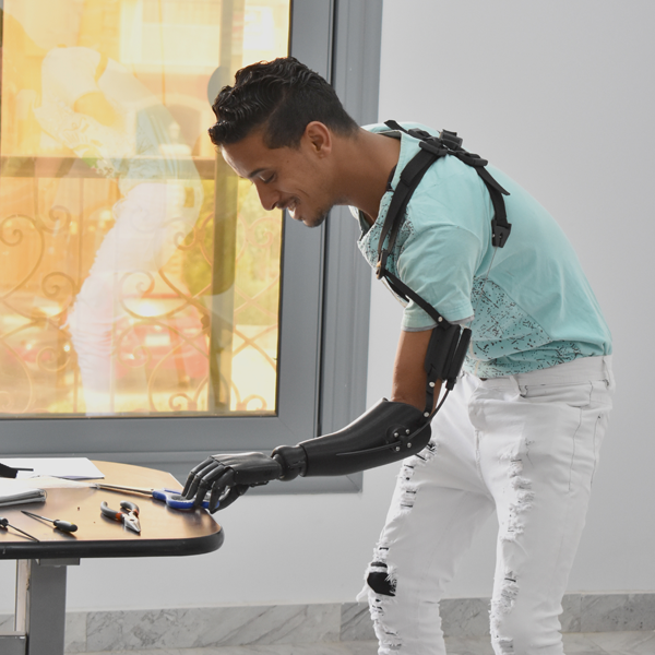 A young man of Color with short hair and a turquoise t-shirt wears a black prosthetic arm and reaches for a pair of scissors lying on a table.
